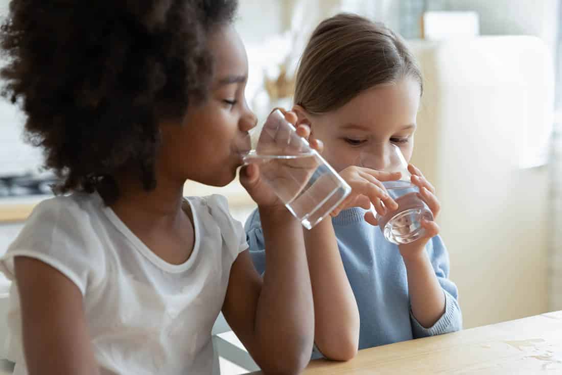 Two multiracial girls sit in kitchen feels thirsty drink water Two girls sitting in kitchen drinking purified water